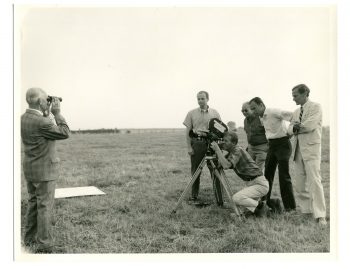 A photo depicting a group of five men huddled around a camera (left) with William Gaddis (far right), all working on the U.S. army film, The Battle of St. Vith.