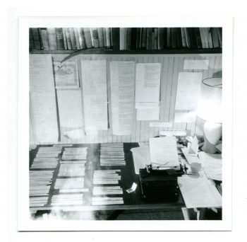 A photo of William Gaddis's workroom. The desk and wall are covered with intricately laid out, typed papers; there are books on shelves in the very top of the photo frame; and a typewriter with a lamp rests on the right of the desk. 