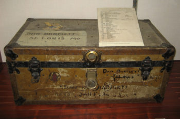 The image displays an worn footlocker, or luggage trunk. The trunk has "Dan Bartlett St Louis MO" written on the lid, along with a number of other, worn scribbles along its front. 