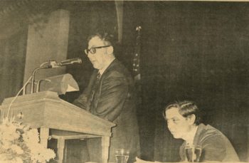 A photo of Stanley Elkin speaking at a podium while William Gass, also on-stage, looks on.