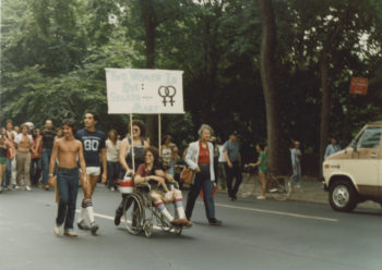 Groups of people walking down the middle of the street during the Sea Cliff Pride Parade. In the foreground of the photo are five individuals, four walking abreast of each other and one in a wheelchair. A sign attached to the wheelchair with poles is mostly illegible, but does have two female gender symbols side-by-side.