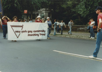 Groups of people walking down the middle of the street holding a banner during the Sea Cliff Pride Parade. The banner reads "New York Gay Community Marching Band."