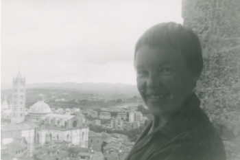 May Swenson smiling at the camera with an expanse of Siena, Italy, in the background. 