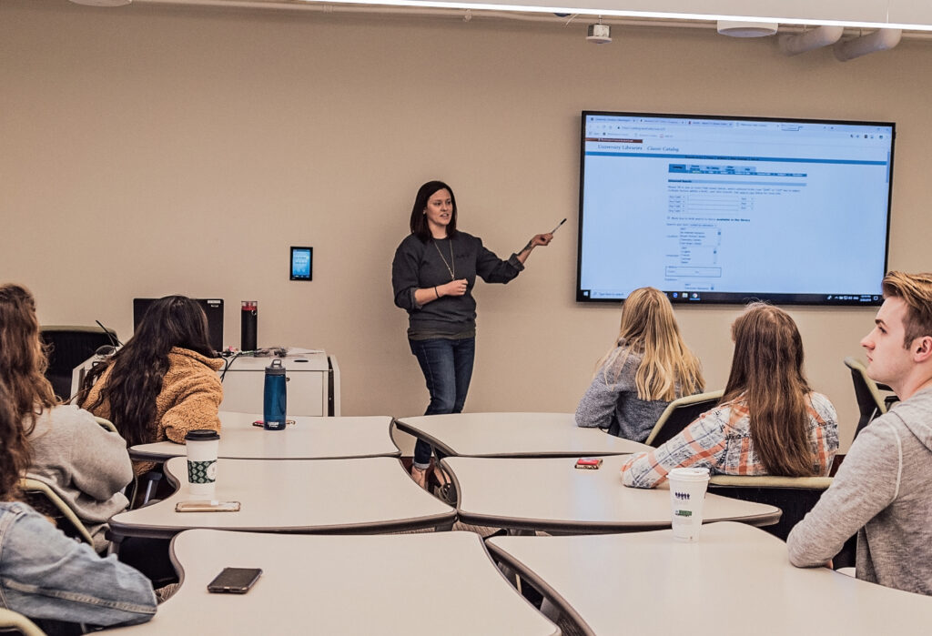 Amanda Albert is standing in front of a large digital screen showing instruction notes while a mix of six students sit listening. 