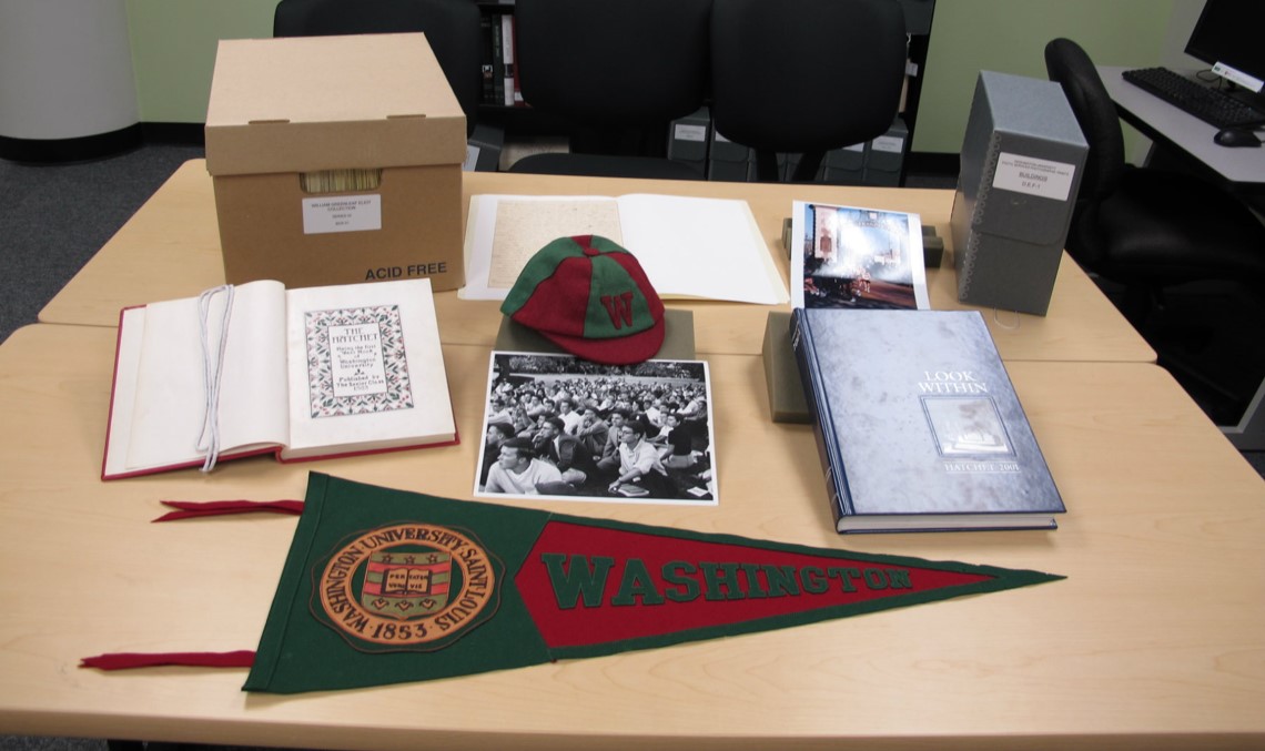 A small collection box with its contents displayed on the table. The Collection consists of Washington University items from the past - there is an older flag and sports cap in the WashU colors; a text open to a highly stylized bookplate with a placeholder ribbon; a thicker, leather-bound text; some photographs; a glossy pamphlet; and a smaller collection box that typically holds papers or lose documents. 