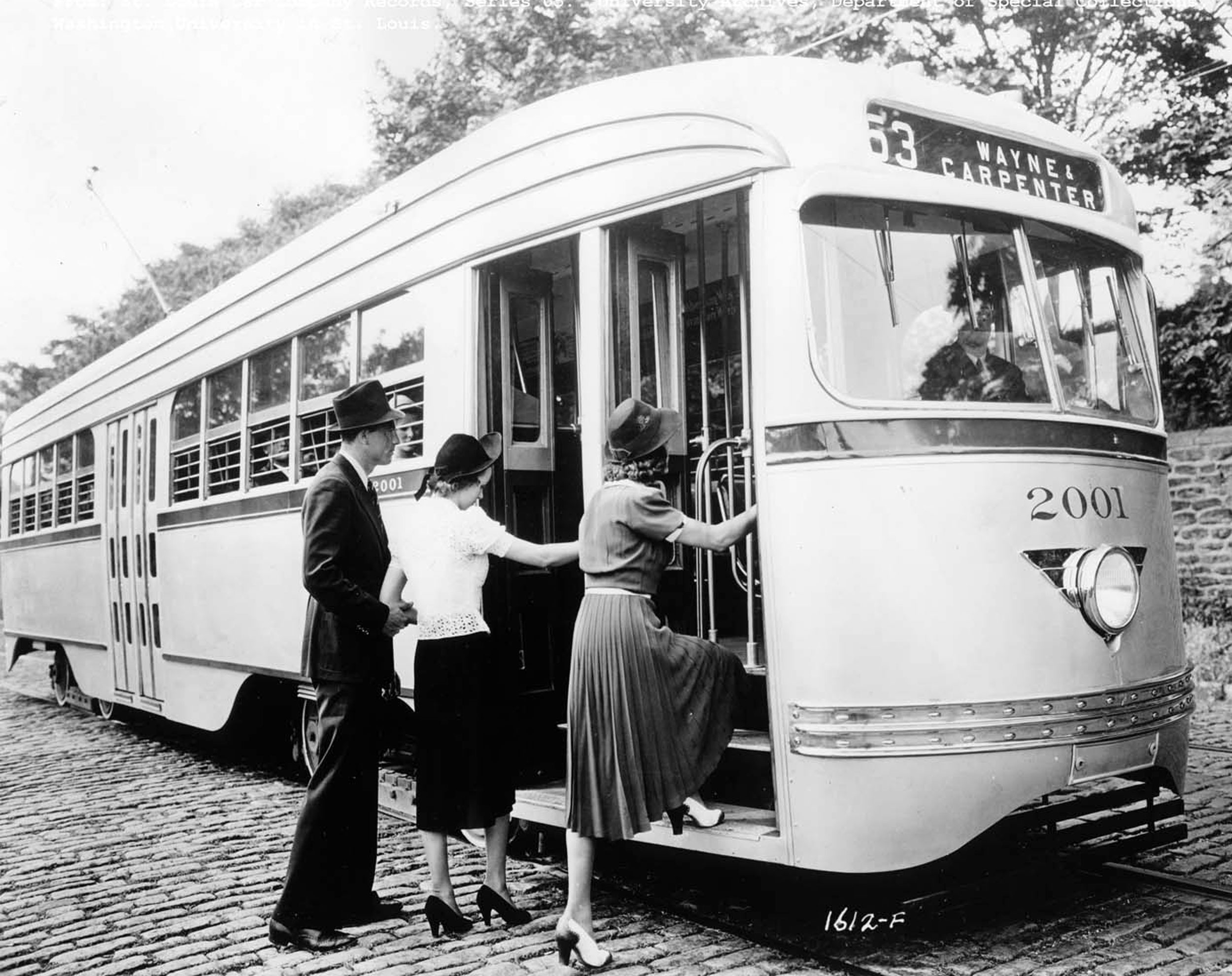 A black and white photo of a gentleman in a top hat helping two ladies off the cobblestone street and onto the Wayne & Carpenter #63 Streetcar in Saint Louis.