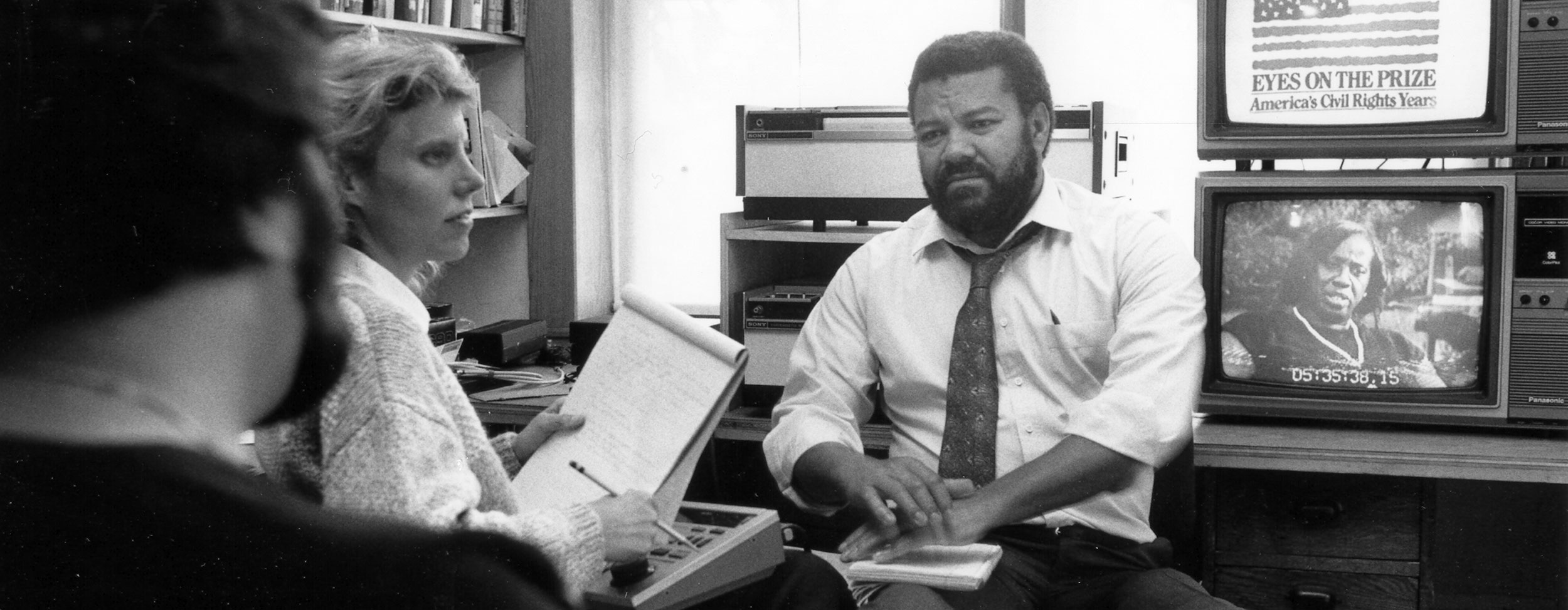 Henry Hampton with Blackside colleagues Kenn Rabin and Cindy Kuhn. Kuhn looks to be taking copious notes while Hampton and Rabin discuss a topic. Eyes on the Prize and an interview play on two TVs in the background.