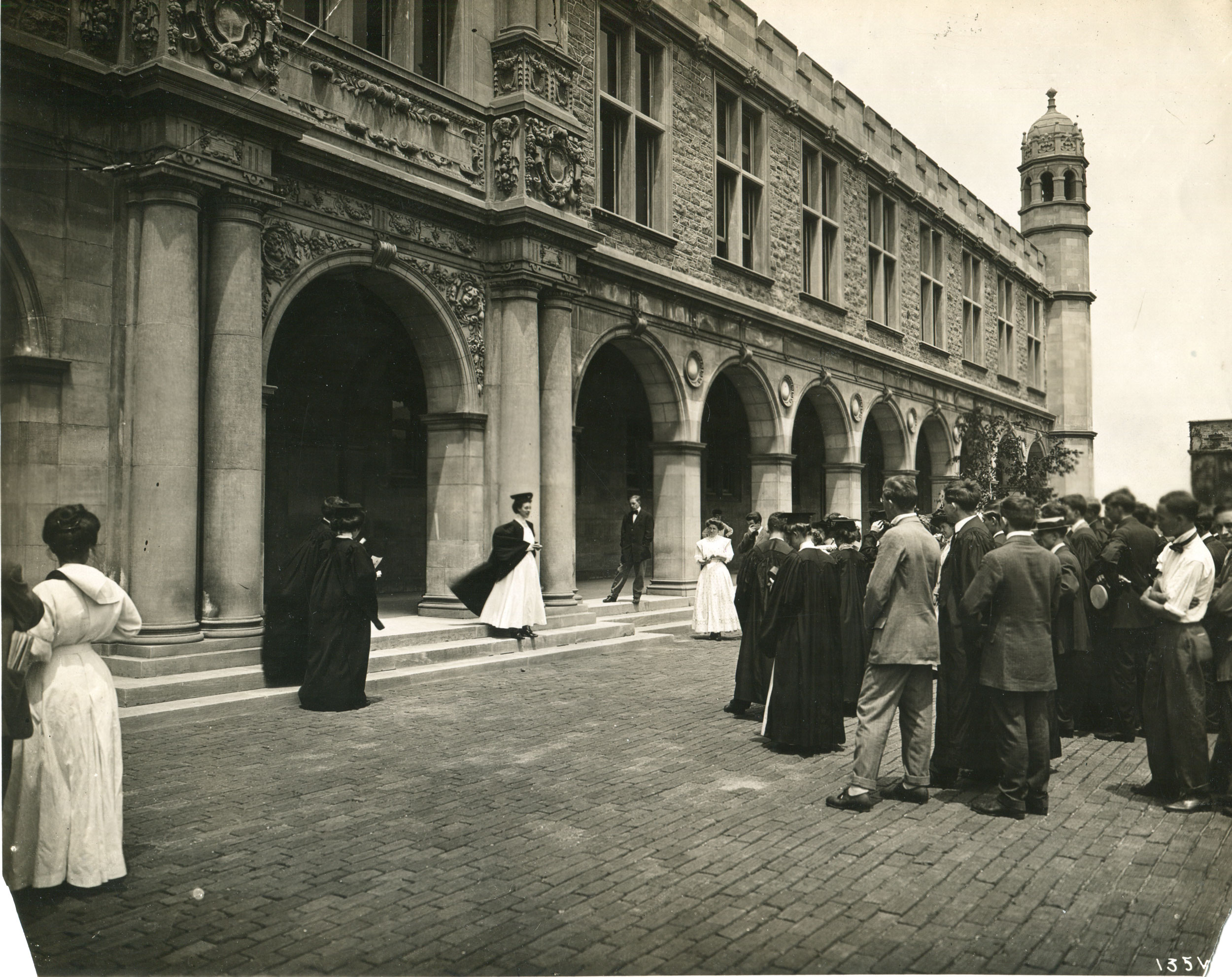 The photo shows a group of graduates and others celebrating commencement. A female student stands at the center beneath the archway entrance to Ridgely Hall being photographed in a white gown and commencement robes.