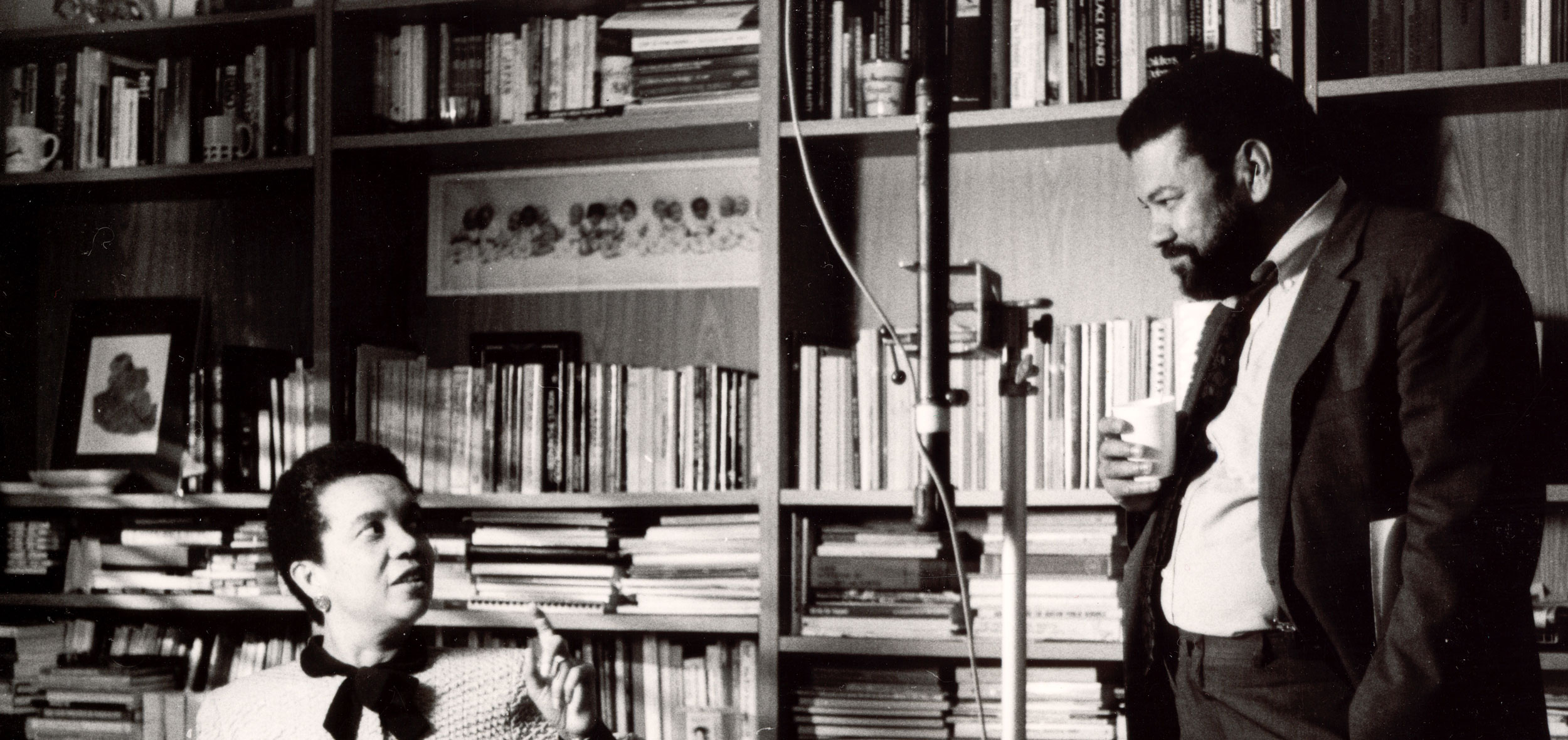 Filmmaker Henry Hampton stands in front of a heavily occupied bookshelf with a seated Marian Wright Edelman nearby.