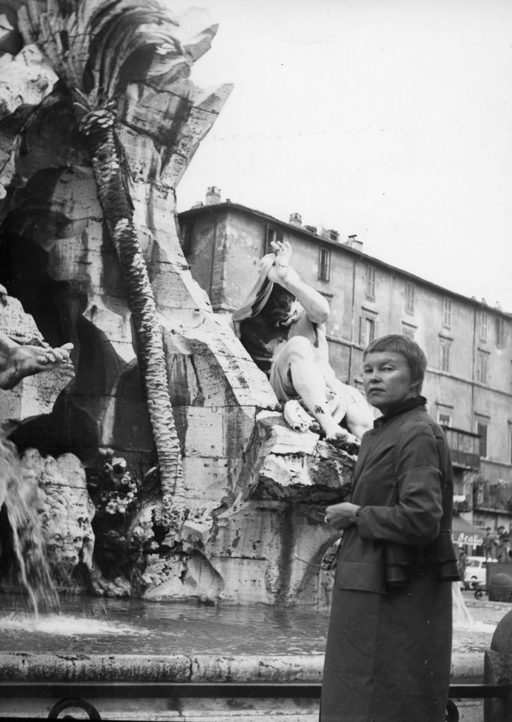 May Swenson stands in front of a fountain wearing a long coat.