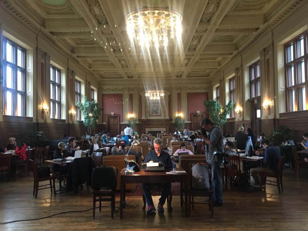 A pjhoto of Youd sitting at a wooden desk with a lamp and typewriter in the midst of a crowded Holmes Lounge being photographed retyping The Franchiser. 