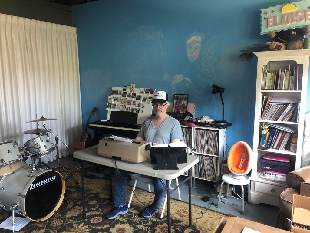 Youd sitting at a collapsable table with a typewriter retyping The Tunnel in his garage/studio. The Garage has a curtain partition set up, along with a rug on the floor, bookshelves, and a drum set, amongst other things. 