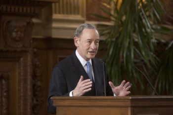 Chancellor Mark S. Wrighton standing in a suit and tie at a podium softly gesticulating with his hands during his opening remarks at Gass's memorial service. 