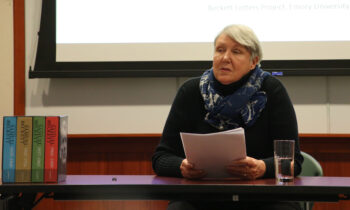 Lois Overbeck sitting at a long, panel table presenting from a collection of papers held in their hands. 