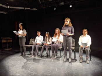 Six students with six chairs in a row performing "The Conscious Inexistence" on a spotlight lit stage. The first and fifth students are standing while reading from scripts in their hands. 