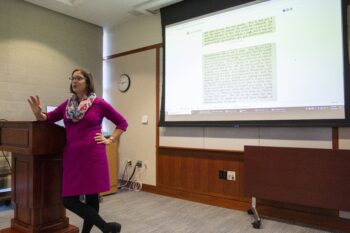 Anna Teekell presenting at What is the World: Celebrating Samuel Beckett. Teekell stands at the front of the room to the side of a podium in front of a projection screen displaying talking points. 