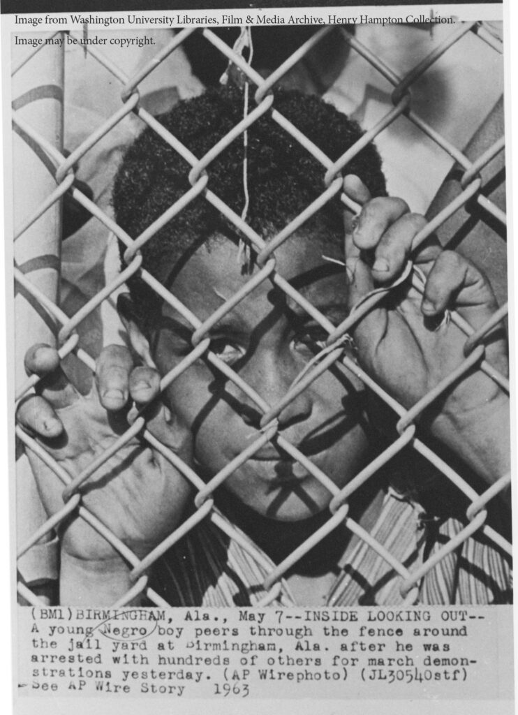 Young boy behind a chainlink fence. The caption reads "Birmingham, Ala., May 7. Inside Looking Out. A young Negro boy peers through the fence around the jail yard at Birmingham, Ama., after he was arrested with hundreds of others for march demonstrations yesterday."