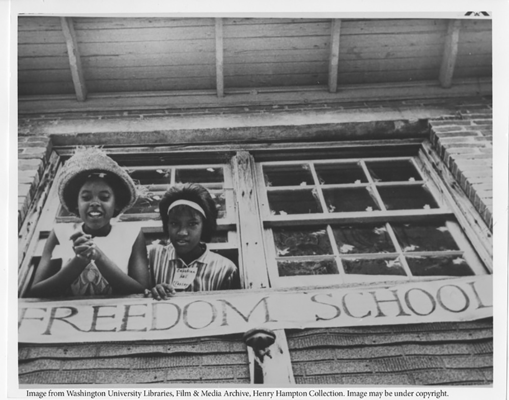 Two young girls with their hair nicely done and in pressed tops peer out of a double window with a banner beneath them reading "Freedom School."