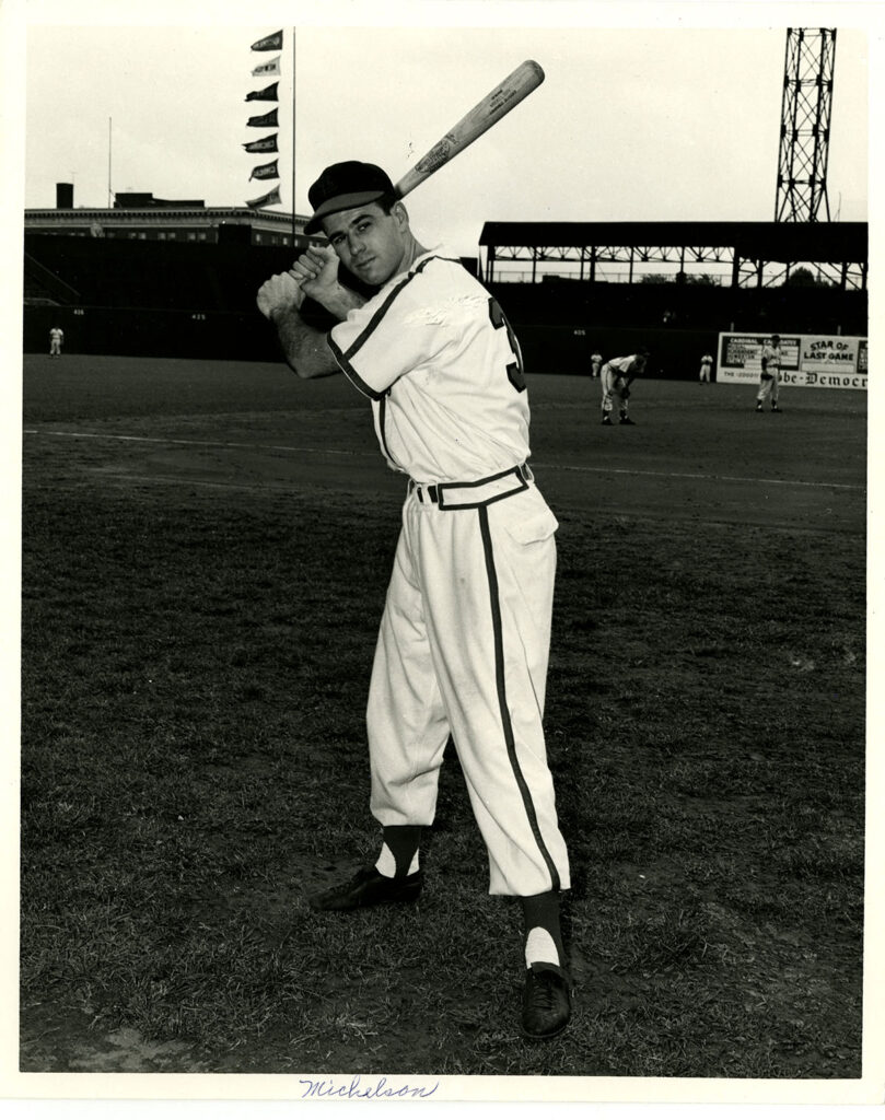 Mickelson is in-uniform on a baseball field with a ball-cap and is photographed with a bat ready to swing.