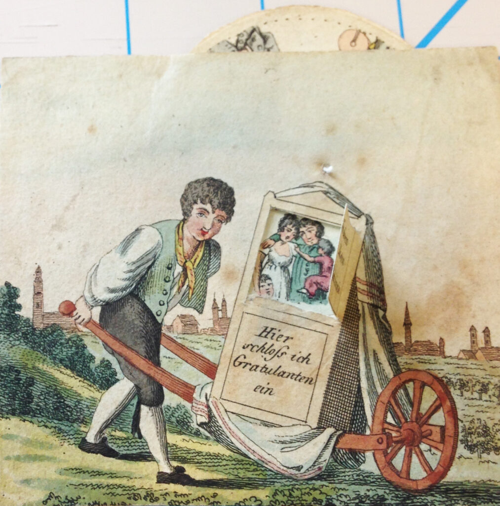 A curly haired child with a vest over a long, puffed-sleeved shirt wearing a knotted handkerchief around his neck with britches and fancier shotes totes a puppet theater box about on a single-wheeled wheelbarrow.