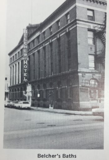 A very grand building in appearance. Four-stories with a high ceiling in the ground floor. There is a sign with the Hotel's name (Belcher Bath Hotel) along the front and hints of a fire escape just out of frame. 