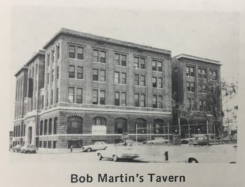 A large, decorated four-story building. The ground-floor has high, arched entrances; there are eight windows across the building facing the photographer with six windows along the corner. 