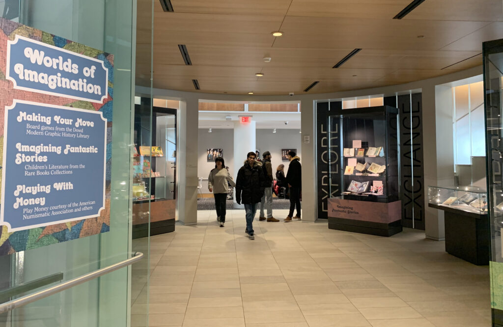 A photo of the Worlds of Imagination exhibition with students investigating the materials. A Worlds of Imagination promotional sign describes the three parts of this exhibition: Making Your Move, Board games from the Dowd Modern Graphic History Library; Imagining Fantastic Stories, children's literature from the Rare Books Collection; and Playing with Money, play money courtesy of the American Numismatic Association.