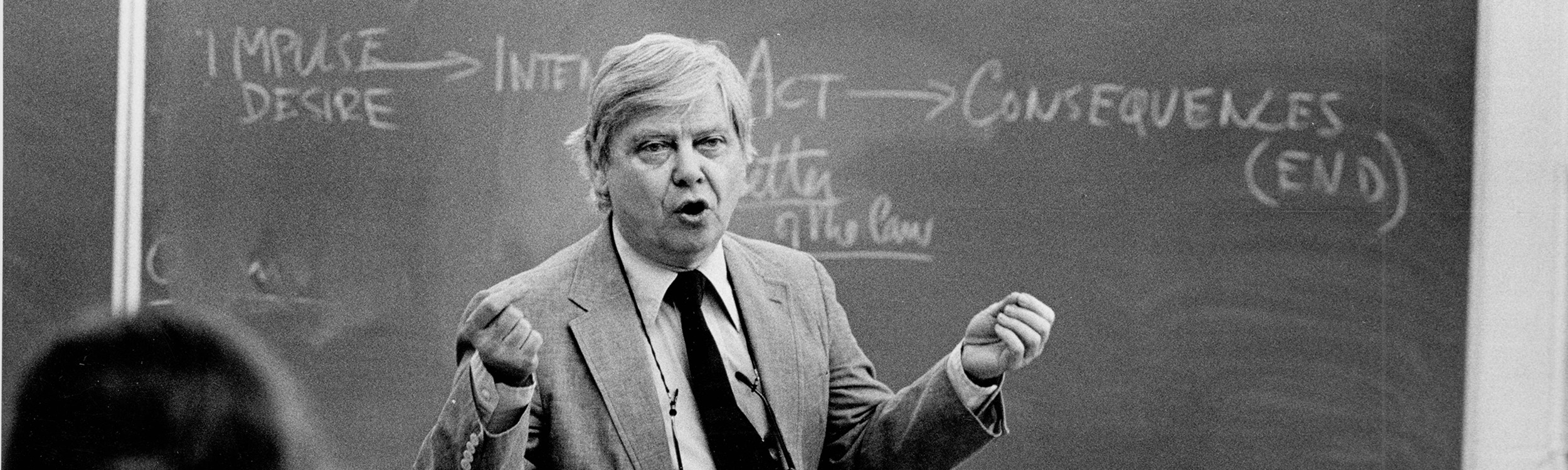 Gass stands in front of a chalkboard in a suit and tie. The photograph has captured him mid-speech during a lecture.