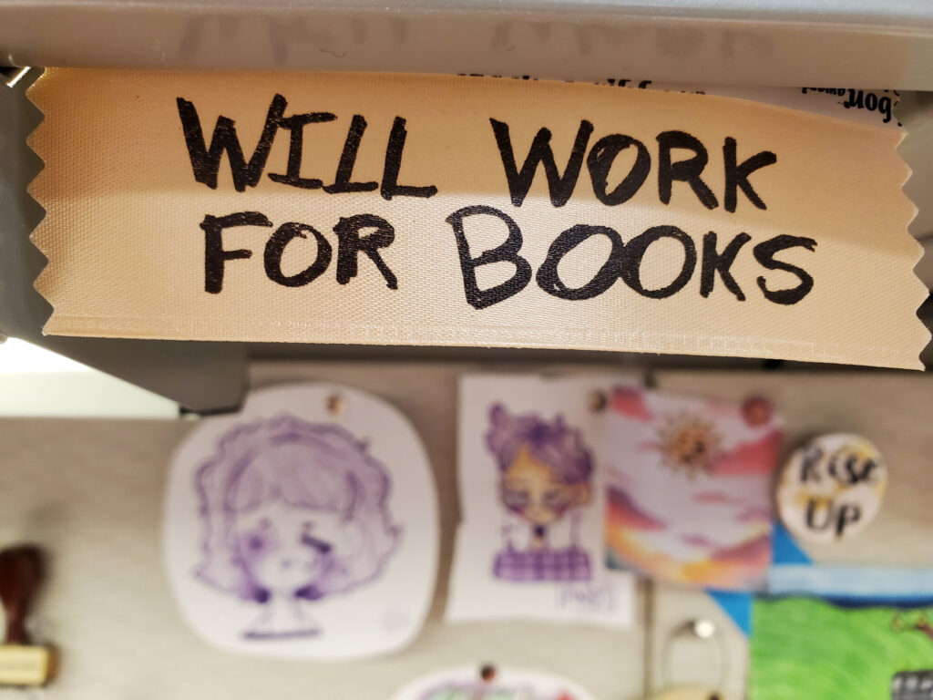 Handwritten decoration on a staff member's desk that reads "will work for books" in all-caps.