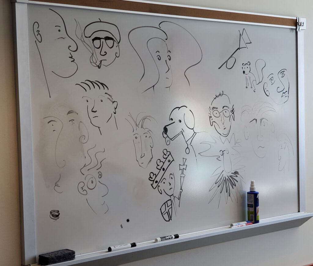 Doodles done on a white board in an office. The doodles are mostly of faces, but a dog and a squirrel make an appearance as well.