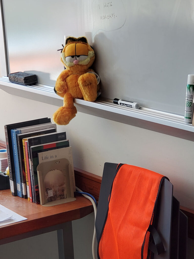 A Garfield the cat stuffed animal lounging on the marker tray of a white board.