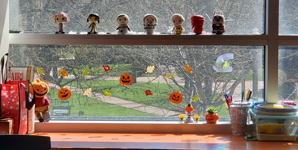 Staff member's desk with Funko Pops and yarn knitted super heros on display. There are Halloween decorations on the window.