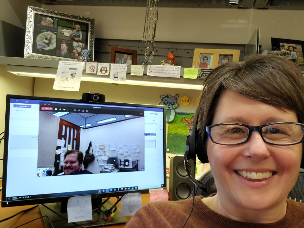 Staff member Ehrstein at their desk with headphones on while a coworker, Brady, is displayed in a Zoom window on their computer during a virtual meeting.