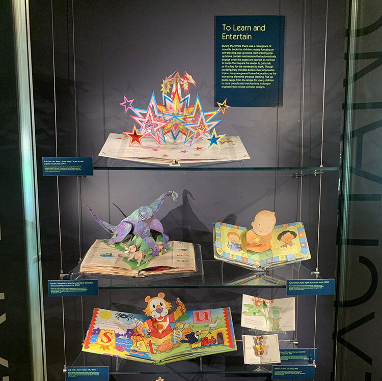 Display titled "To Learn and Entertain," part of the Moving Magic exhibition. The display features eight colorful, educational popup books of various sizes.