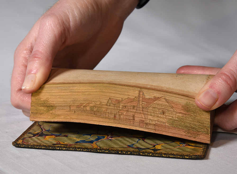 The fore edge is a painting of a village with trees on one side of a lane and row homes on the other. One can also see the stone fence and chimney of the closest house.