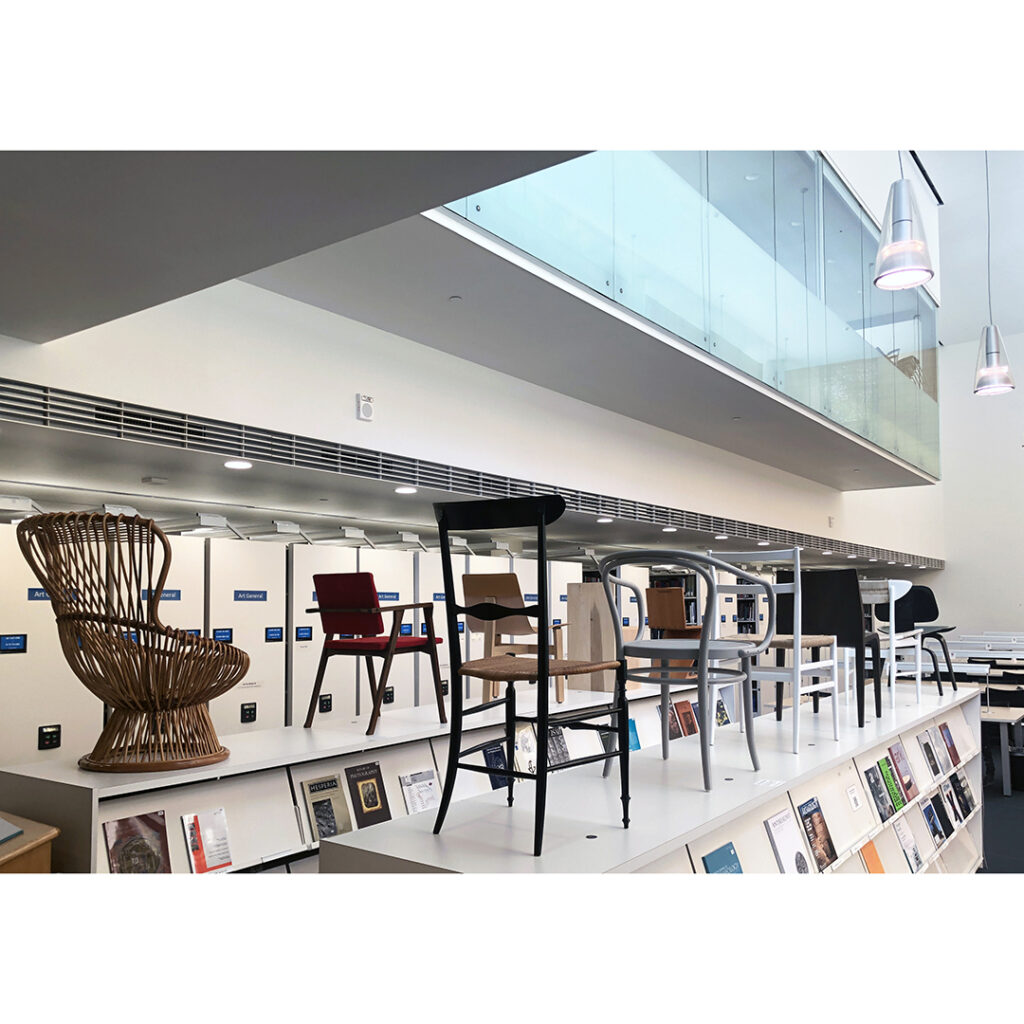 The photo is of the exhibition, chairs of various design on top of magazine shelves inside the Art & Architecture Library.