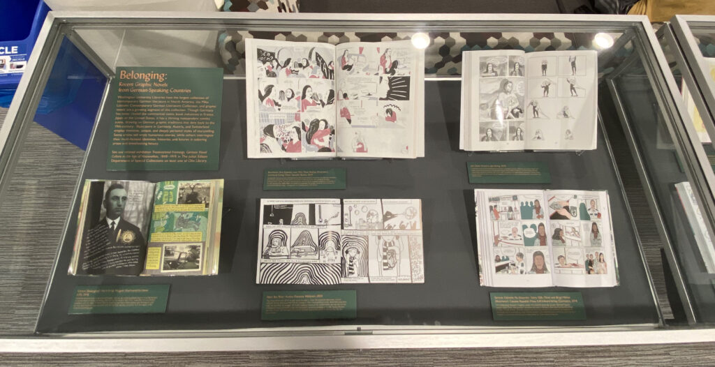 The image shows a display case with five graphic novels open in two-page displays. Each graphic novel has a placard alongside it explaining the individual texts.