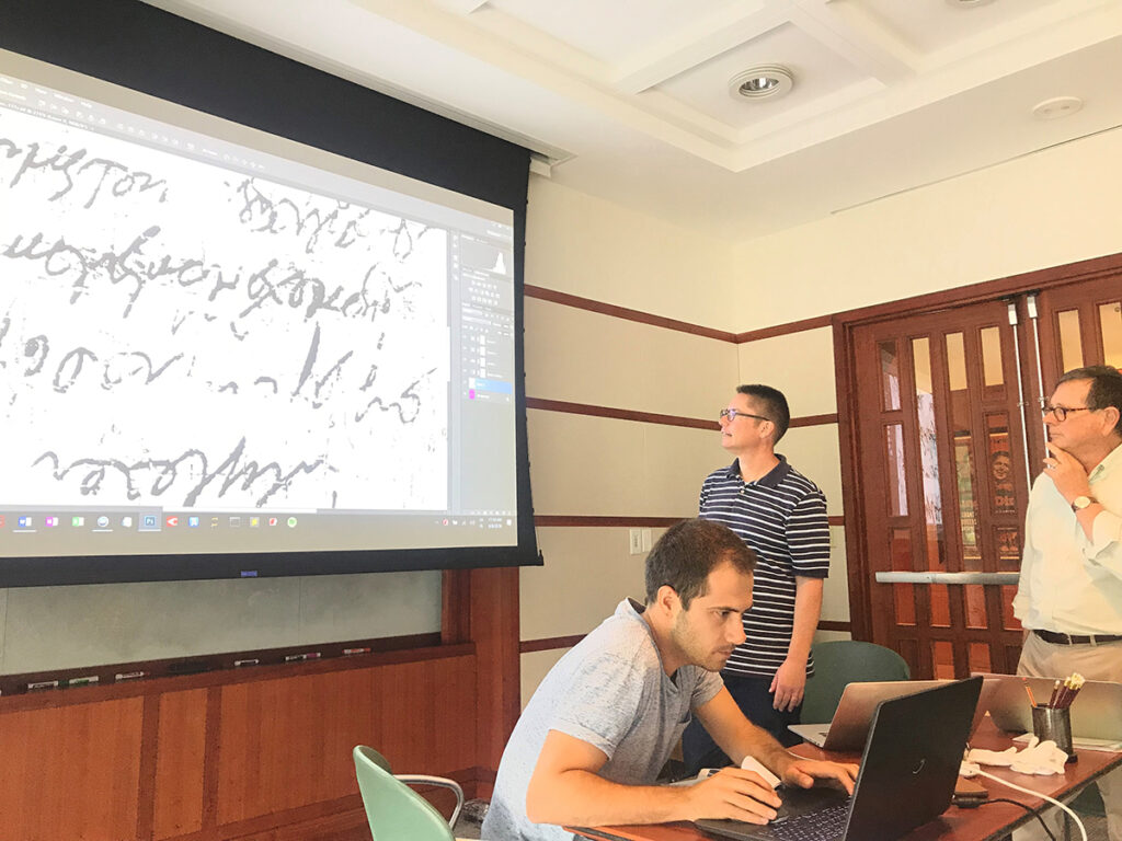 Three individuals (two standing, one seated) study a section of scanned papyri that is projected onto a white board.