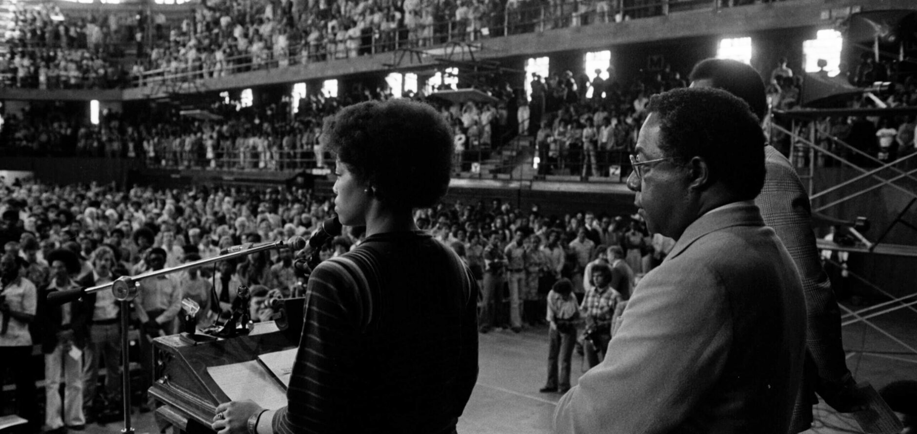 Jan Taylor, president of the Association of Black Student’s program committee, stands speaking at a podium in front of a crowded Field House facility while introducing Assembly Series speaker Alex Haley in 1977.