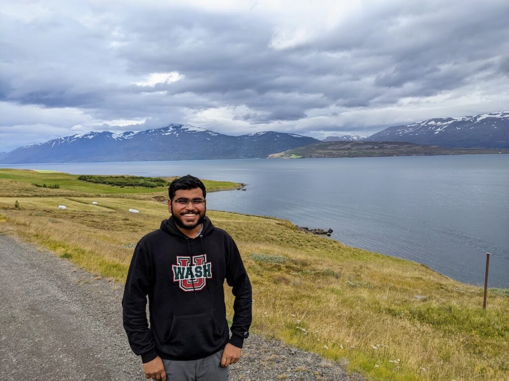 Person standing in front of a fjord with snow-capped hills beyond the fjord.