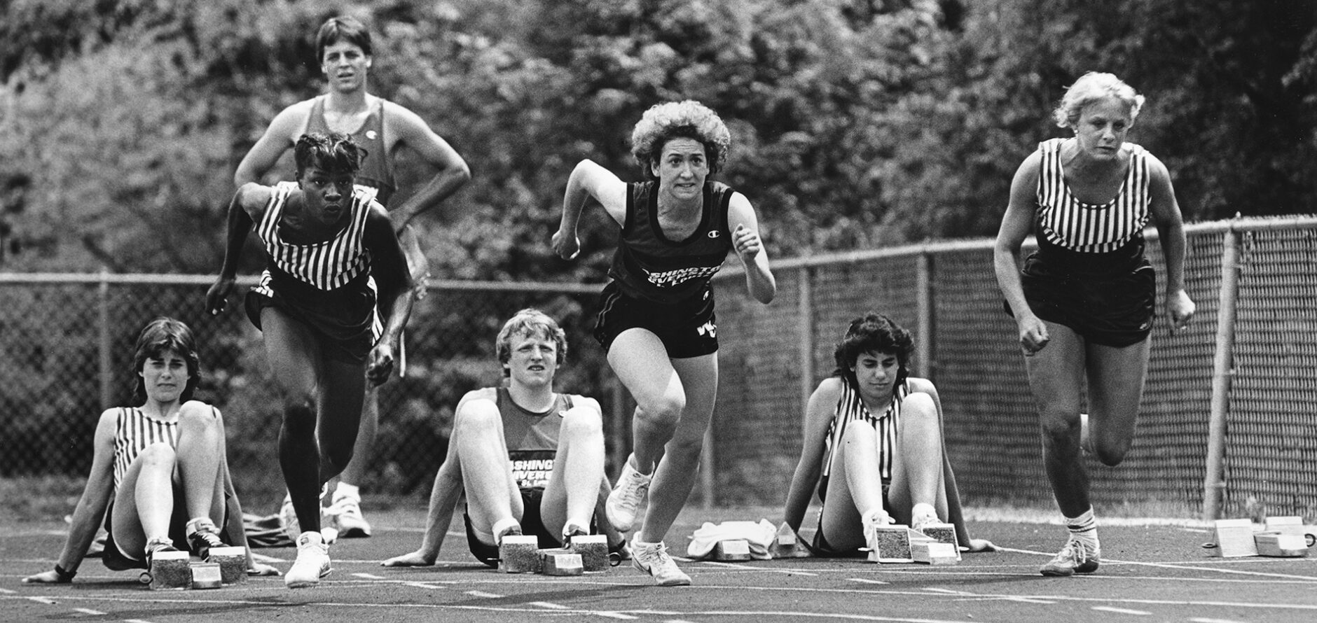 Three female track runners leaving the block as four of their teammates watch.