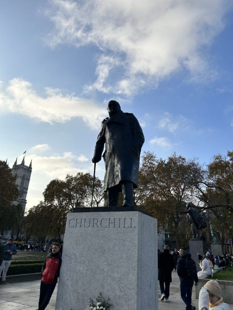 A statue with a walking stick mounted on a tall pedestal with the word 'Churchill' in a public park.