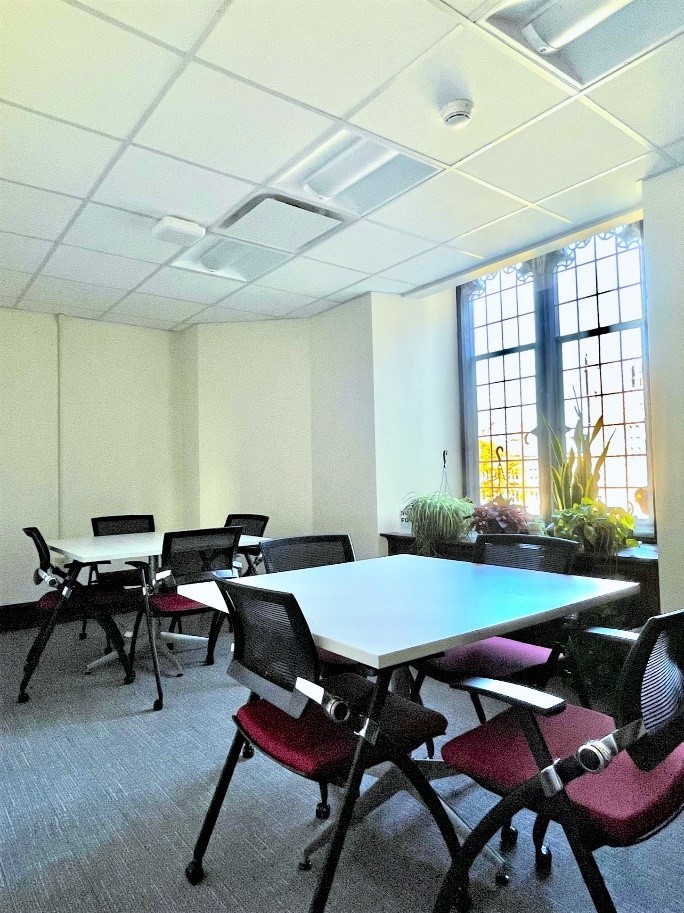 A window pane with potted plants overlooking a room with two tables each with four chairs.