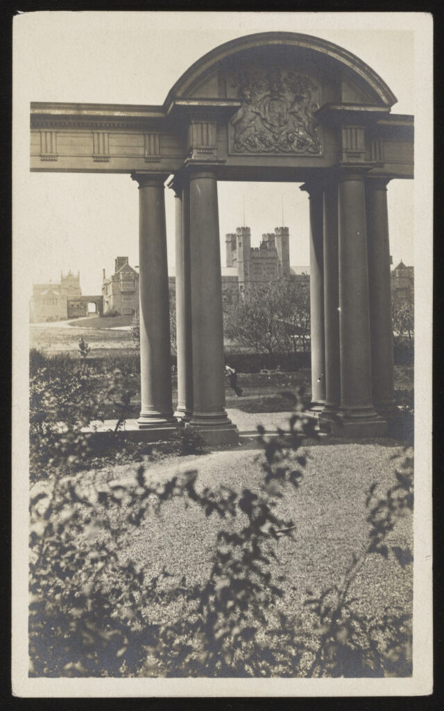 An archway with columns and an arched roof with a garden around it and campus buildings in the background.