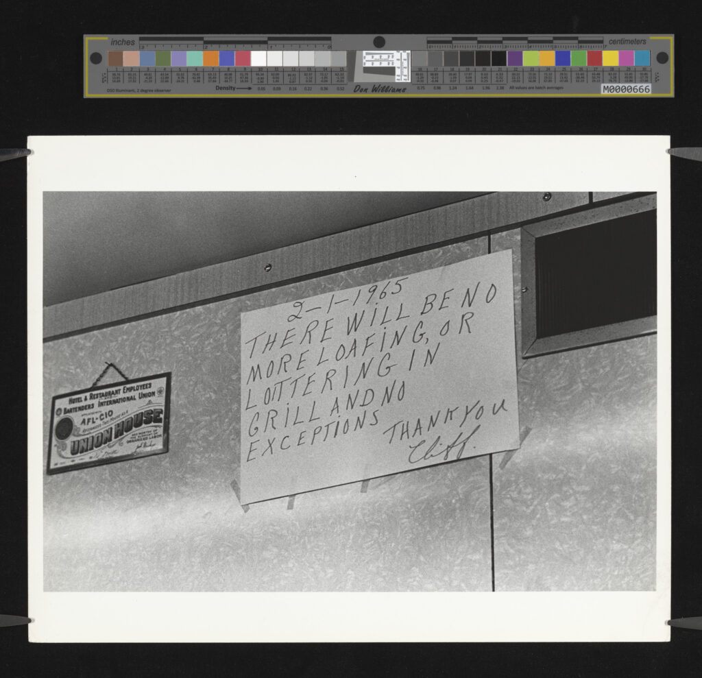 A black and white photo of a picture and a hand-written sign dated 2-1-1965 hanging and taped to a wall respectively