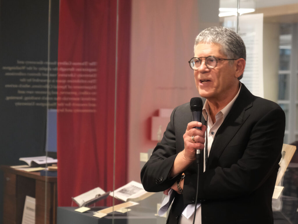 A man speaking into a mic in front of an exhibit behind glass cases.