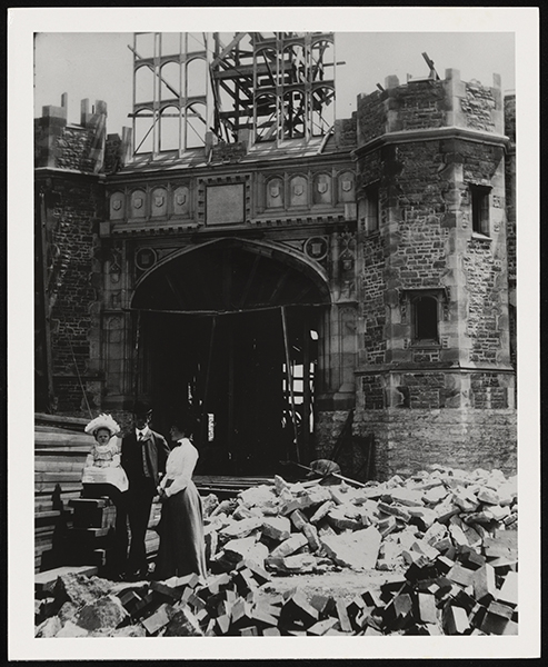 Man, woman, and child in fancy clothes and hats stand among the rubble and lumber at the entrance of a partically constructed Brookings Hall.