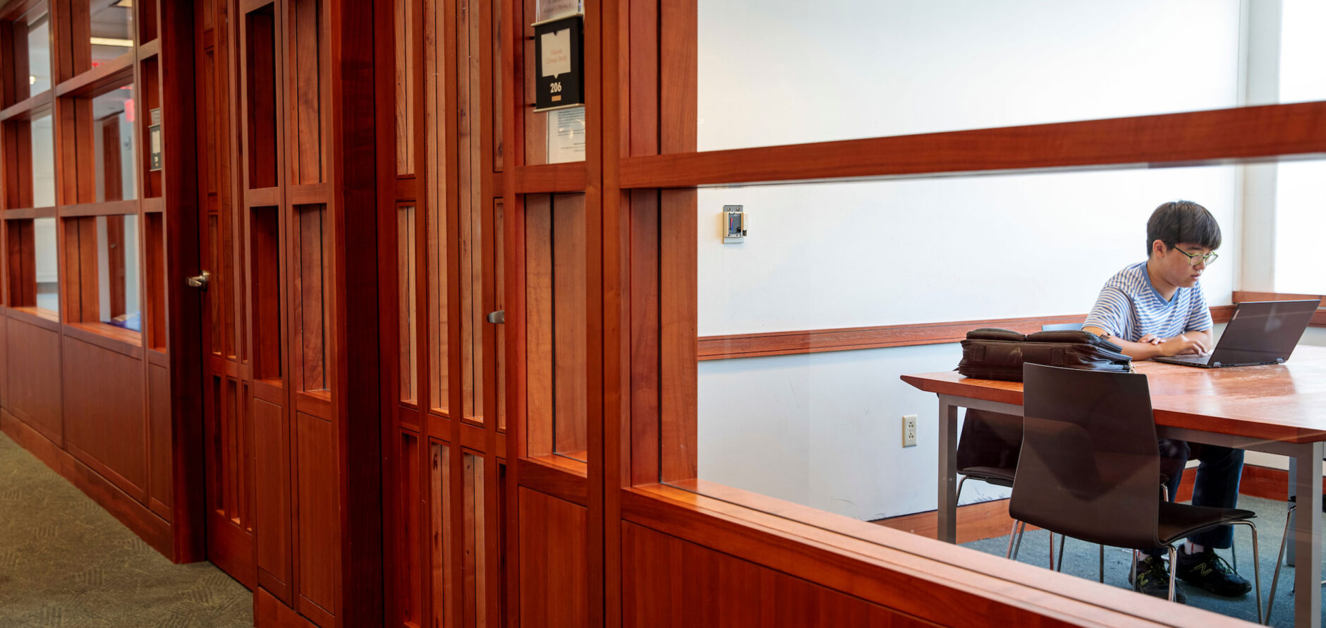 A view of a study room from the hall outside. The room can be seen through a large window and there is a student seated at the table inside.