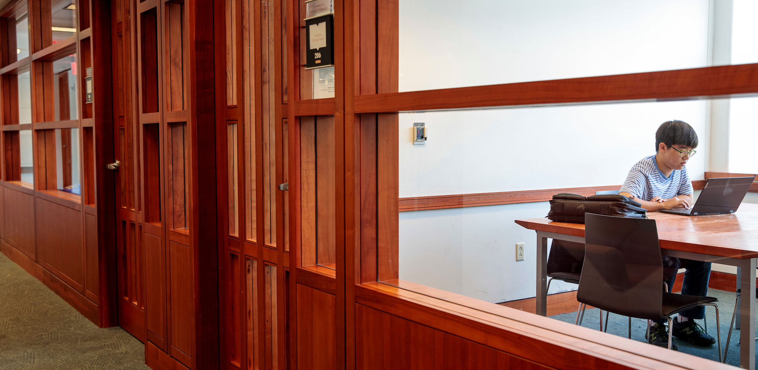 A view of a study room from the hall outside. The room can be seen through a large window and there is a student seated at the table inside.