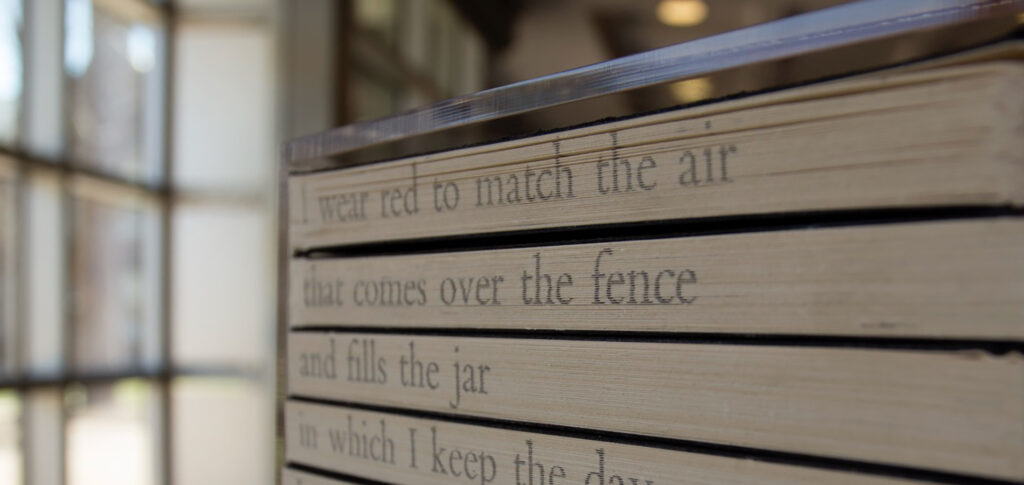 A sculpture of spine-in books on a shelf with the beginnings of a poem written on the pages that reads: I wear red to match the air / that comes over the fence / and fills the jar / in which I keep the day...